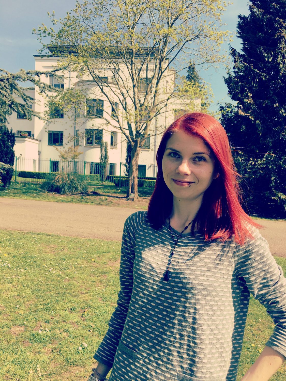 Caz (Young, white woman with red hair) smiling while standing in the park during a sunny day.